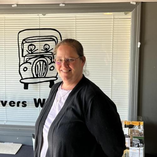 smiling woman in grey and black standing in front of a window with the two men and a truck logo