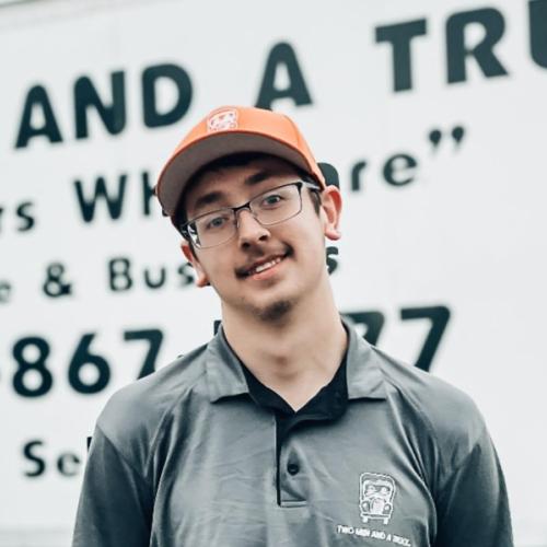 Kaylob is wearing an orange hat and he is standing outside in front of moving trucks