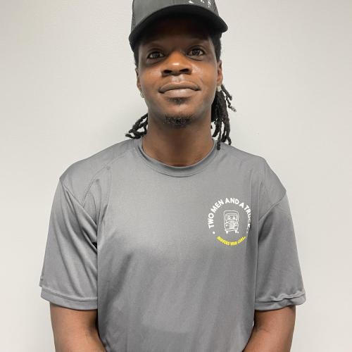 headshot of a smiling man standing in front of a grey wall wearing a grey two men and a truck tshirt
