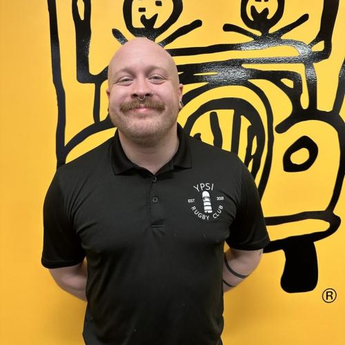 john is wearing all black standing in front of a yellow wall with the truckie logo