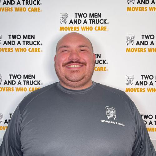 miguel sanchez standing in front of two men and a truck background wearing grey shirt