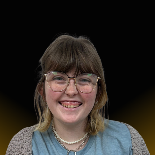 smiling woman wearing glasses, two necklaces and a blue tshirt with grey sweater