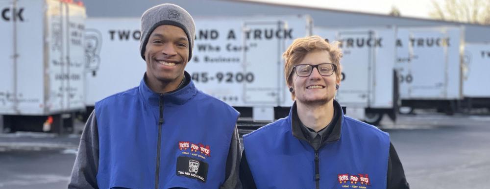 Two men standing in front of the trucks wearing a blue Toys for Tots vest