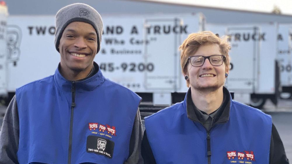 Two men standing in front of the trucks wearing a blue Toys for Tots vest