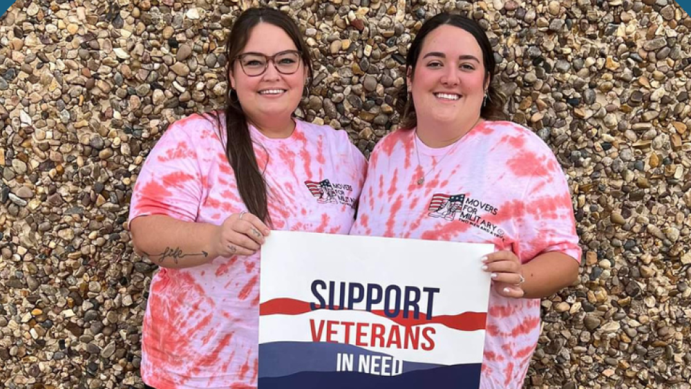 two women standing in front of rock wall wearing red tie dye shirts holding sign for veterans