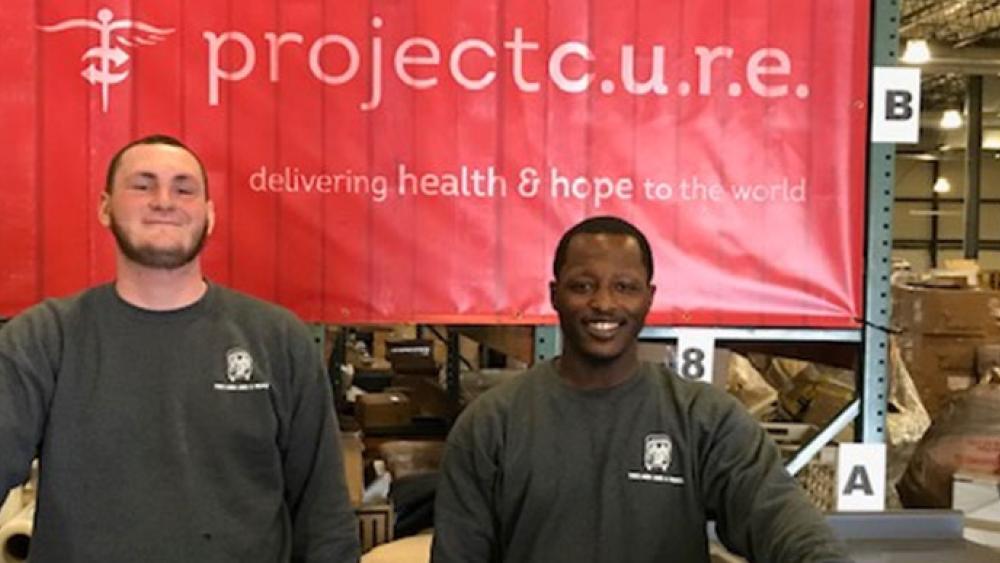 two men and a truck movers standing in front of project cure banner