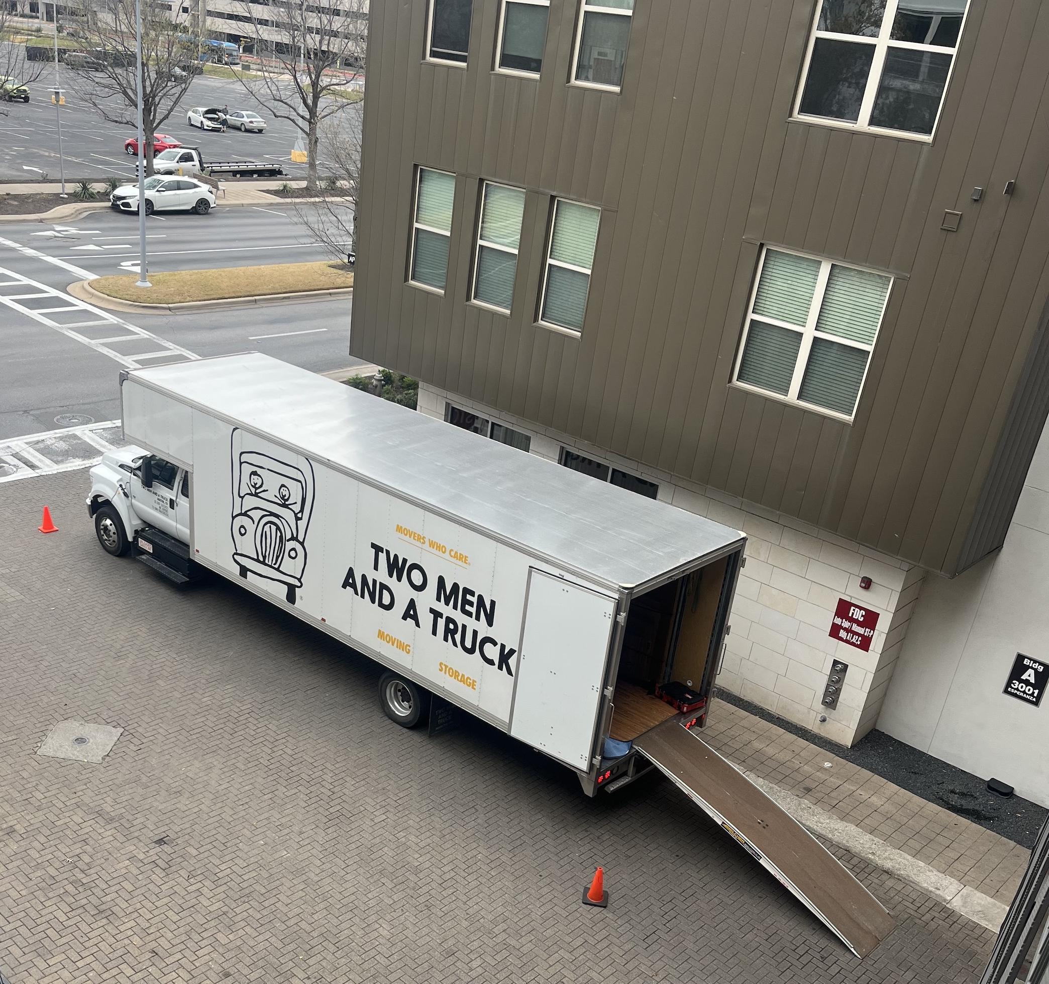 Two Men And A Truck moving truck parked in Austin with the ramp of the out and safety cones in the street. 