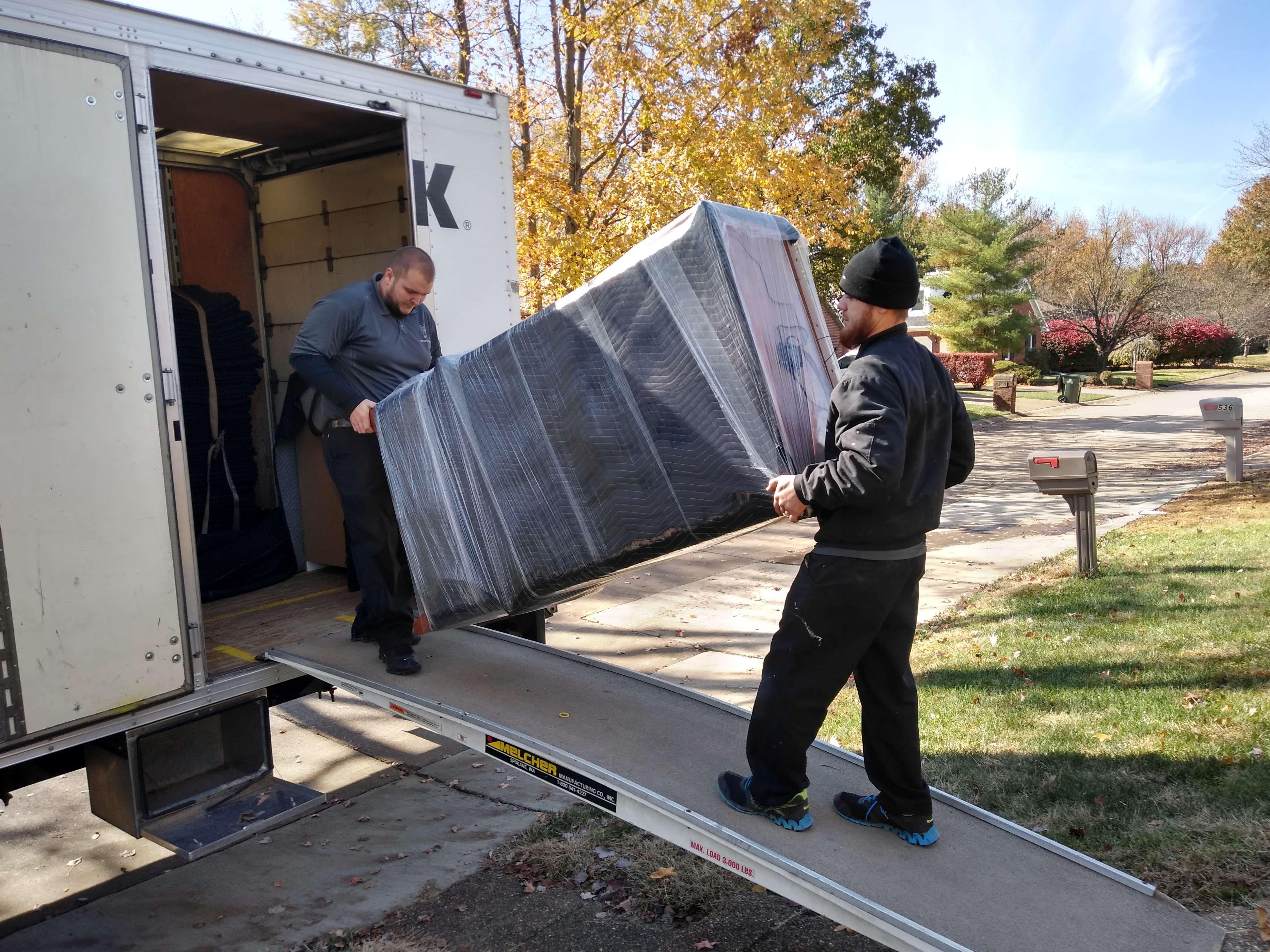 two movers carrying a pad wrapped item up the ramp of the side of the truck