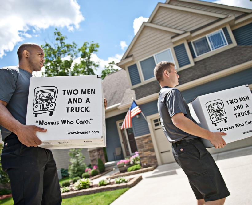 TWO MEN AND A TRUCK movers carry boxes in front of a home.