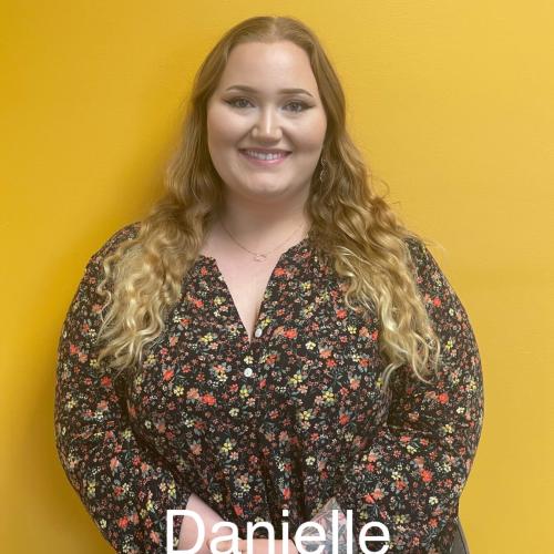 Danielle is wearing a floral shirt, smiling, standing in front of a yellow wall