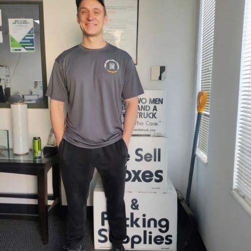 smiling man wearing a baseball cap and a two men and a truck tshirt, standing in front of moving boxes